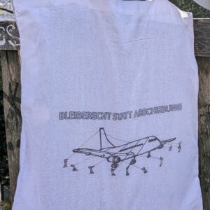 Weiße Tasche mit Aufschrift Bleiberecht statt Abschiebung und festgebundenem Flugzeug