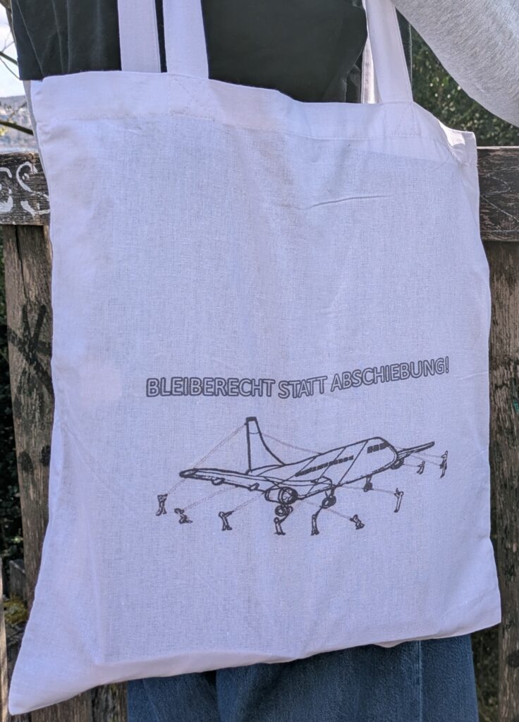 Weiße Tasche mit Aufschrift Bleiberecht statt Abschiebung und festgebundenem Flugzeug